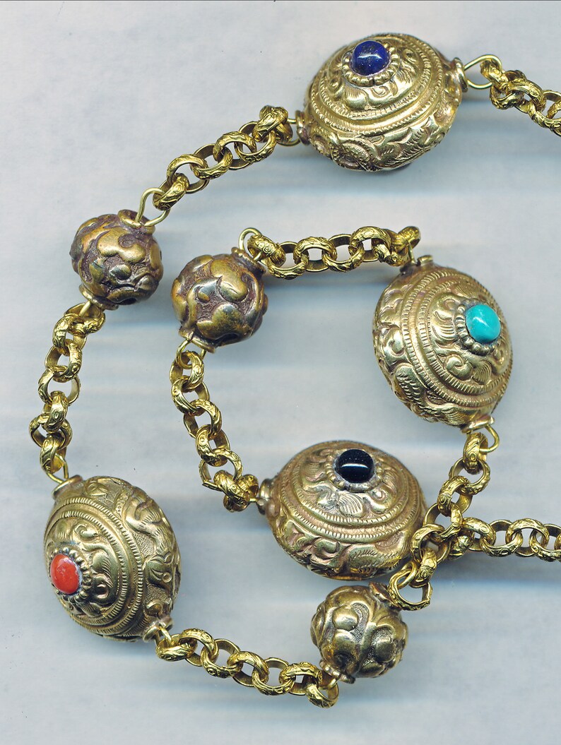 Puede incluir: Un collar de cadena de oro con cuentas redondas y ovaladas. Las cuentas est&aacute;n decoradas con dise&ntilde;os intrincados y engastadas con piedras preciosas azules, turquesa, negras y rojas.