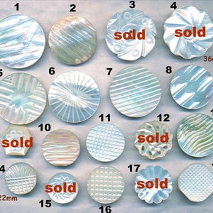 May include: A collection of iridescent mother-of-pearl buttons in various shapes and patterns. Some buttons are round, others are hexagonal or floral. Several have "sold" text. The largest button is 36mm in diameter, and the smallest is 22mm.