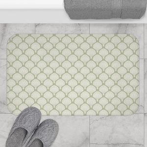 May include: A rectangular bath mat with a repeating fan-shaped pattern in light green and cream. The mat is on a tiled floor, with gray slippers at the bottom and a gray towel at the top.