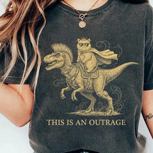 May include: Dark gray t-shirt featuring a cartoon cat riding a dinosaur, wearing sunglasses and a cape. The text "THIS IS AN OUTRAGE" is printed below the graphic. The shirt has a vintage, worn look.