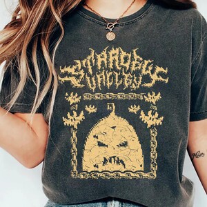 May include: A black t-shirt with a graphic print of a pumpkin with a chain link fence around it. The text "Scareface Hollow" is printed above the pumpkin. The shirt is worn by a person with long blonde hair.