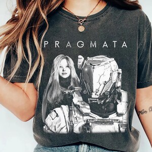 May include: Dark gray t-shirt with the word "PRAGMATA" printed across the top. The shirt features a black and white graphic of a woman and a futuristic robot. The shirt is a vintage style.