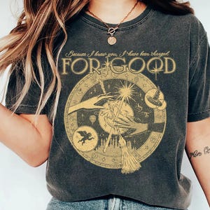 May include: Dark grey t-shirt featuring a gold circular graphic with hands, a star, a crescent moon, and a broom. The text "Because I knew you, I have been changed FOR GOOD" is printed above the graphic in gold.