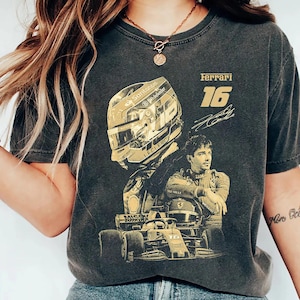 Könnte beinhalten: Dunkelgraues T-Shirt mit einem goldfarbenen Aufdruck eines Formel-1-Rennwagens, eines Fahrers und eines Helms. Das Shirt zeigt das Wort "Ferrari" und die Nummer "16".