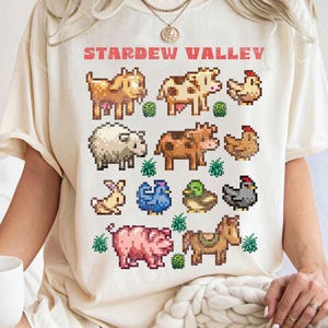 Op de afbeelding: Crèmekleurig T-shirt met gepixelde boerderijdieren en de tekst "Stardew Valley" in het rood. Het ontwerp bevat koeien, kippen, schapen, een konijn, een varken, een paard en andere boerderijdieren. Het shirt is een casual, comfortabele stijl.