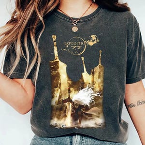Może przedstawiać: Ciemnoszara koszulka z złotą grafiką i napisem "Expedition 73". Projekt zawiera postać z mieczem i stylizowany krajobraz. Koszulka ma wygląd vintage.
