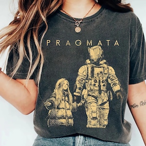 Könnte beinhalten: Dunkelgraues T-Shirt mit dem Wort "PRAGMATA" oben aufgedruckt. Das Shirt zeigt eine goldfarbene Illustration einer futuristischen Astronautenfigur, die die Hand eines jungen Mädchens hält. Das Mädchen trägt einen Puffmantel.