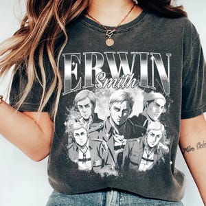 May include: Dark grey t-shirt with the name "ERWIN Smith" in a silver, vintage-style font. Below the text are illustrations of five anime characters. The shirt has a vintage appearance.