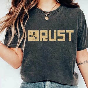 May include: Dark grey t-shirt featuring the word "RUST" in a distressed, tan font. The shirt has a crew neck and short sleeves. The text is centred on the chest. The shirt is worn with blue jeans.
