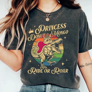 Puede incluir: Camiseta gris oscuro con un dinosaurio de dibujos animados montado por un gato con corona y capa, sobre un c&iacute;rculo arco&iacute;ris. El texto dice "Princess Donut Mongo" arriba y "Ride or Roar" debajo del gr&aacute;fico.