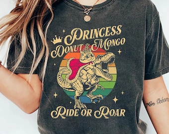 May include: Dark gray t-shirt featuring a cartoon dinosaur ridden by a cat wearing a crown and cape, set against a rainbow circle. The text reads "Princess Donut Mongo" above and "Ride or Roar" below the graphic.
