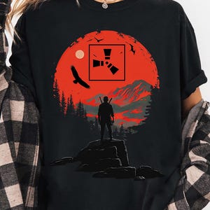 May include: Black t-shirt with a graphic of a person on a rock, set against a red sunset. The design includes a square logo, an eagle, trees, and mountains. The shirt is suitable for casual wear.