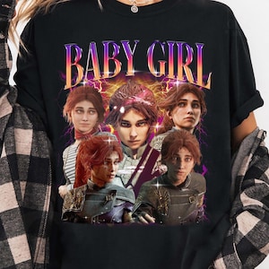 May include: Black long-sleeved t-shirt with a graphic design. The design features the text "BABY GIRL" in a purple and orange gradient font, and multiple portraits of a person with red hair.