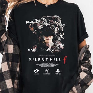 May include: Black t-shirt featuring the text "SILENT HILL f" with a graphic of a person with a floral headpiece. The shirt also includes the text "RETURN TO SURVIVAL HORROR" and logos for KONAMI, N00BARDS, and PS5.