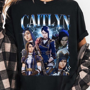 May include: Black t-shirt with a graphic print of a group of characters from the video game "League of Legends". The characters are Caitlyn, Vi, Jinx, and Jayce. The text "CAITLYN" is printed in blue and white letters above the characters.