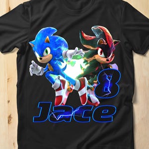 Könnte beinhalten: Schwarzes T-Shirt mit Sonic the Hedgehog und Shadow the Hedgehog. Die Zahl 8 und der Name "Jace" sind in blauem Neon-Text dargestellt. Sonic ist blau mit weißen Handschuhen und rot-weißen Schuhen. Shadow ist schwarz und rot.