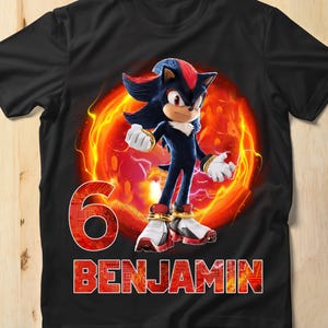 Könnte beinhalten: Schwarzes T-Shirt mit einer roten und orangenen Grafik von Shadow the Hedgehog aus der Sonic the Hedgehog Videospielserie. Die Grafik enthält auch die Zahl "6" und den Namen "BENJAMIN".