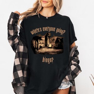 Może przedstawiać: Czarny t-shirt z sepiową grafiką przedstawiającą scenę wioski i napisem "where's everyone going? bingo?". Projekt przedstawia budynki, ognisko i wieżę. Koszulka jest noszona z koszulą w kratę.