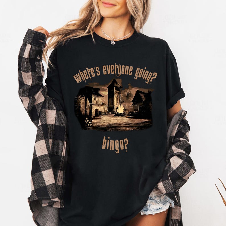 Może przedstawiać: Czarny t-shirt z sepiową grafiką przedstawiającą scenę wioski i napisem "where's everyone going? bingo?". Projekt przedstawia budynki, ognisko i wieżę. Koszulka jest noszona z koszulą w kratę.
