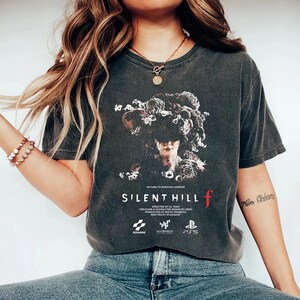 May include: Dark gray t-shirt featuring a graphic from "Silent Hill f." The design includes a detailed illustration of a figure with floral elements, the game's title, and the logos of Konami, NeoBards, and PS5. The text "Return to Survival Horror" is also visible.