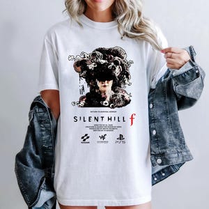May include: White t-shirt featuring a graphic from the video game Silent Hill f. The design includes a detailed illustration of a figure with a dark, ornate headpiece, the game's title, and logos.