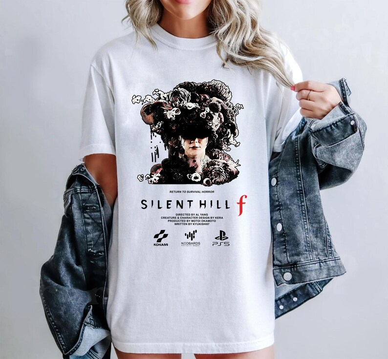 May include: White t-shirt featuring a graphic from the video game Silent Hill f. The design includes a detailed illustration of a figure with a dark, ornate headpiece, the game's title, and logos.