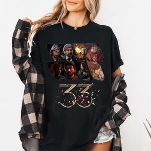 May include: Black t-shirt featuring a graphic design with multiple character portraits and the number "33" in gold. The design includes red and gold confetti-like accents. The shirt is worn by a person.