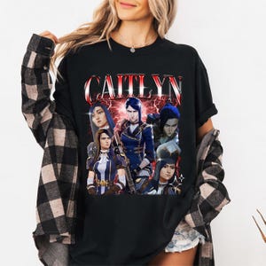 May include: Black t-shirt with a graphic print of a group of characters from the video game League of Legends. The text "Caitlyn" is printed in large, bold letters above the characters.