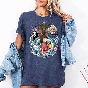Op de afbeelding: Een marineblauw t-shirt met een kleurrijke afbeelding uit de animatiefilm Spirited Away. Het ontwerp bevat personages als Chihiro, No-Face en Yubaba, met een draak, gebouw en kersenbloesems.