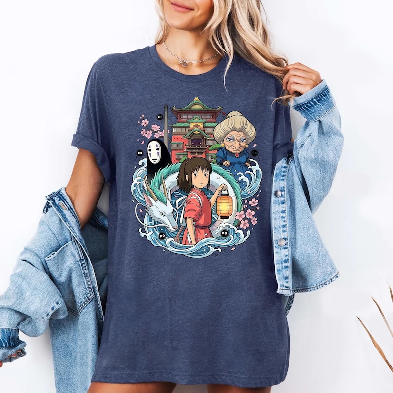 Op de afbeelding: Een marineblauw t-shirt met een kleurrijke afbeelding uit de animatiefilm Spirited Away. Het ontwerp bevat personages als Chihiro, No-Face en Yubaba, met een draak, gebouw en kersenbloesems.