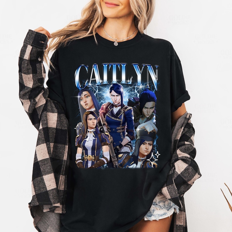 May include: Black t-shirt with a graphic print of a group of characters from the video game "League of Legends". The characters are Caitlyn, Vi, Jinx, and Jayce. The text "CAITLYN" is printed in blue and white letters above the characters.
