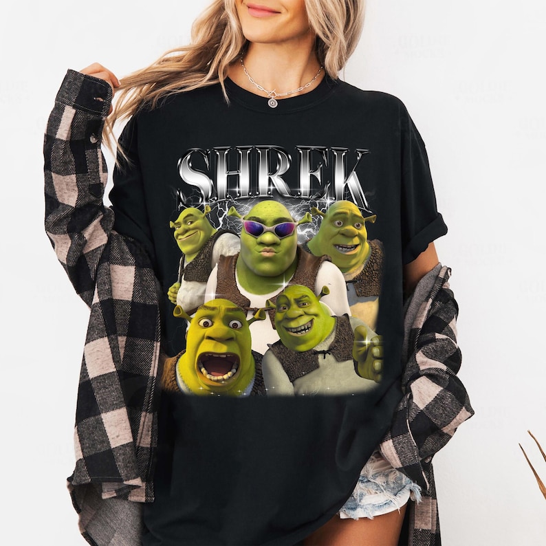 May include: Black t-shirt with a graphic print of Shrek and other characters from the Shrek movie franchise. The text "SHREK" is printed in silver letters on the shirt.