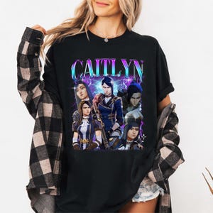 May include: Black t-shirt with a graphic print of a group of characters from the video game League of Legends. The text "CAITLYN" is printed in a large, bold font above the characters.