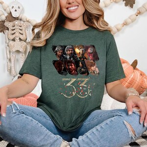 May include: Dark green t-shirt featuring a collage of character images and the number "33" in gold. The design includes a variety of faces and figures, with a festive confetti-like pattern around the numbers.