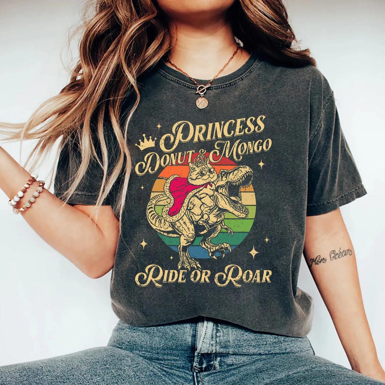 Puede incluir: Camiseta gris oscuro con un dinosaurio de dibujos animados montado por un gato con corona y capa, sobre un c&iacute;rculo arco&iacute;ris. El texto dice "Princess Donut Mongo" arriba y "Ride or Roar" debajo del gr&aacute;fico.