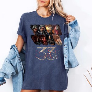 May include: Navy blue t-shirt featuring a collage of character portraits and the number "33" in gold with red confetti. The shirt has a relaxed fit and short sleeves. The design is printed on the front.
