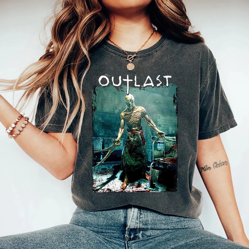Puede incluir: Camiseta gris oscuro con la palabra "OUTLAST" sobre un gr&aacute;fico de un personaje de terror sosteniendo instrumentos quir&uacute;rgicos. El personaje est&aacute; en un entorno m&eacute;dico, con una mesa de operaciones ensangrentada en el fondo.