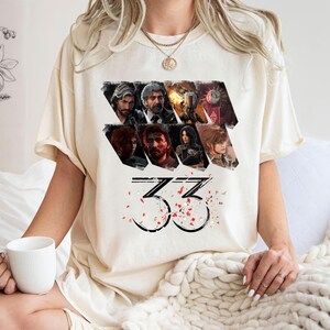 May include: Cream-colored t-shirt featuring a graphic design with multiple character portraits and the number "33" in a stylized font. Red and white confetti-like shapes are scattered around the number. The shirt is made of a soft material.