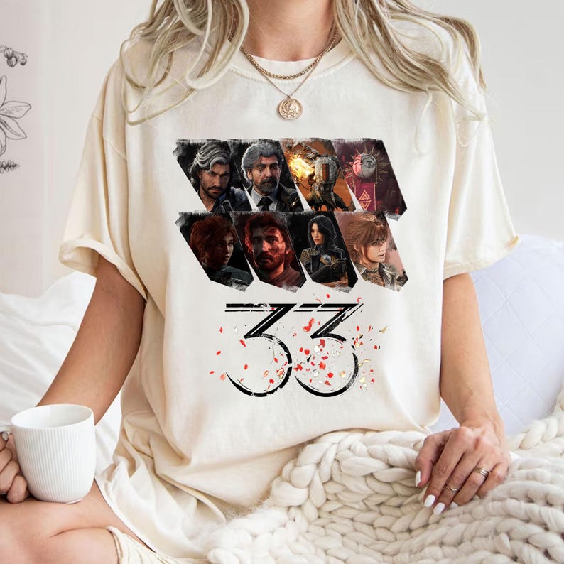 May include: Cream-colored t-shirt featuring a graphic design with multiple character portraits and the number "33" in a stylized font. Red and white confetti-like shapes are scattered around the number. The shirt is made of a soft material.