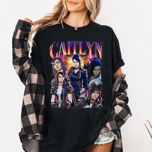 May include: Black t-shirt with a graphic print of a group of characters from the video game League of Legends. The text "Caitlyn" is printed in large, colorful letters above the characters.