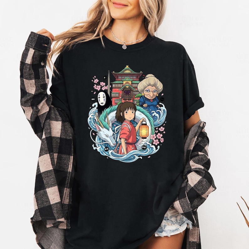 Op de afbeelding: Zwart t-shirt met een kleurrijke illustratie uit de film Spirited Away. Het ontwerp toont personages als Chihiro, No-Face en Yubaba, samen met een draak en Japanse gebouwen. Het kunstwerk is omgeven door water en kersenbloesems.