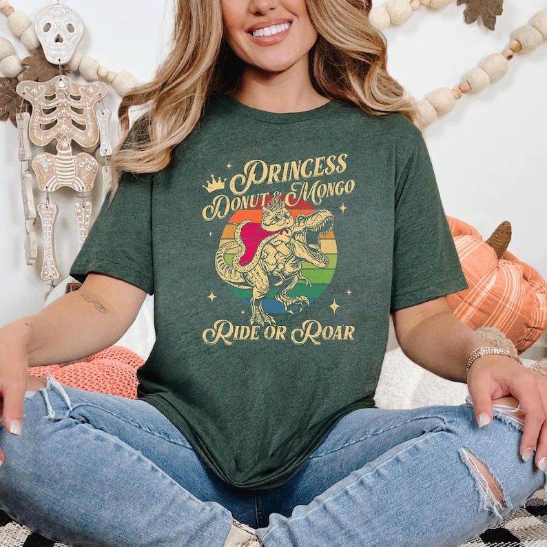 Puede incluir: Camiseta verde oscuro con un dise&ntilde;o de dinosaurio con capa y corona, con el texto "Princess Donut Mongo Ride or Roar". El dise&ntilde;o incluye un c&iacute;rculo arco&iacute;ris y estrellas.