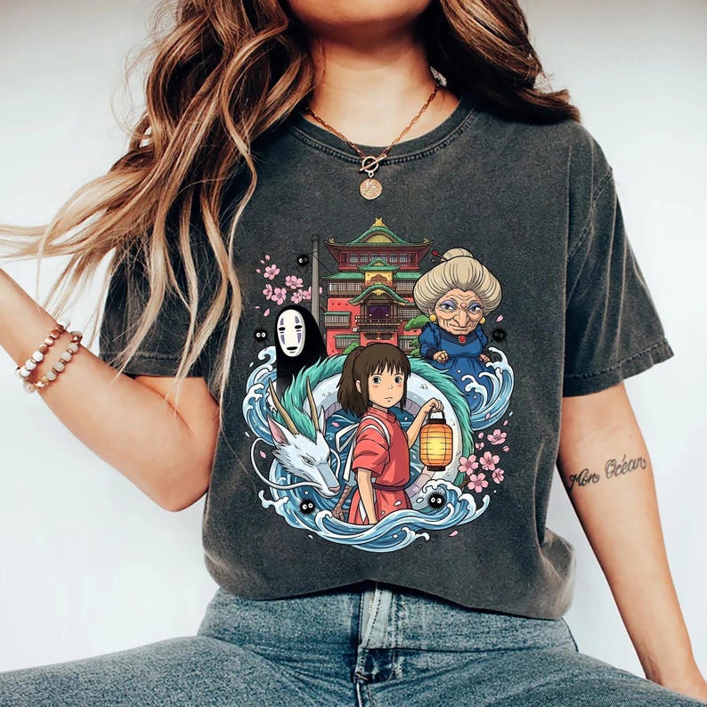 Op de afbeelding: Donkergrijs T-shirt met een kleurrijke anime-stijl grafiek. Het ontwerp bevat een meisje met een lantaarn, een draak en andere personages, tegen een Japanse stijl gebouw. Roze kersenbloesems en waterelementen maken het ontwerp compleet.