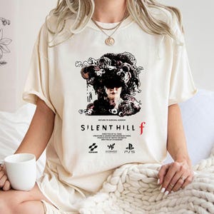 May include: Cream-colored t-shirt featuring the text "SILENT HILL f" in a bold font, with a graphic of a person with a floral headpiece. The shirt also includes the text "RETURN TO SURVIVAL HORROR" and the logos of KONAMI, NEOBARDS, and PS5.