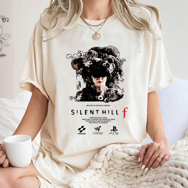 May include: Cream-colored t-shirt featuring the text "SILENT HILL f" in a bold font, with a graphic of a person with a floral headpiece. The shirt also includes the text "RETURN TO SURVIVAL HORROR" and the logos of KONAMI, NEOBARDS, and PS5.