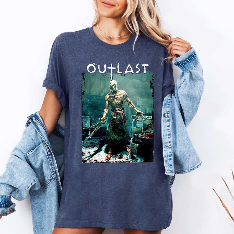 Puede incluir: Camiseta azul marino con la palabra "OUTLAST" en blanco sobre un gr&aacute;fico de una figura amenazante sosteniendo cuchillas. La figura se representa en un entorno oscuro y sangriento.