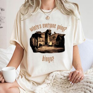 Może przedstawiać: Kremowy t-shirt z sepiowym nadrukiem przedstawiającym scenę miejską i tekstem "where's everyone going? bingo?" wydrukowanym powyżej i poniżej. Koszulka ma kr&oacute;tkie rękawy i luźny kr&oacute;j.