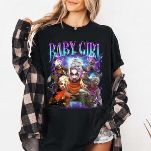 May include: Black t-shirt with a graphic print of a group of cartoon characters. The text "BABY GIRL" is printed in a colorful, retro font above the characters.