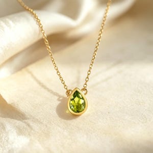 May include: A delicate gold necklace featuring a pear-shaped, light green gemstone pendant. The pendant is set in a gold bezel and hangs from a fine gold chain. The necklace rests on a soft, cream-colored fabric.