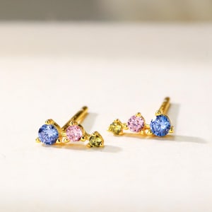May include: A pair of gold stud earrings with three small gemstones. The gemstones are blue, pink, and green.